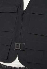 Czarna kamizelka robocza z wieloma przednimi kieszeniami, zamkiem błyskawicznym i zapięciem na zatrzask. Wykonana z lekkiego, teksturowanego materiału.