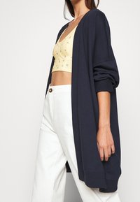 Navyblauer Cardigan aus weichem Stoff, mit offener Vorderseite und langen Ärmeln. Getragen über einem gelben Cropped-Top und weißen Hosen.