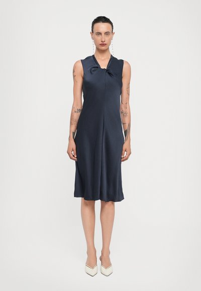 Navy sleeveless dress with a knotted front, made of smooth fabric, featuring a flared hem. Paired with pointed cream heels.