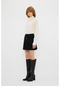 Weißer Rollkragenpullover, schwarzer plissierter Minirock mit goldenen Akzenten und knielange schwarze Leder-Stiletto-Stiefel mit Reißverschluss.