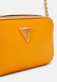 Bolso cruzado amarillo en forma de triángulo hecho de material texturizado. Cuenta con un logo plateado y una correa de cadena, con un cierre con cremallera en la parte superior.