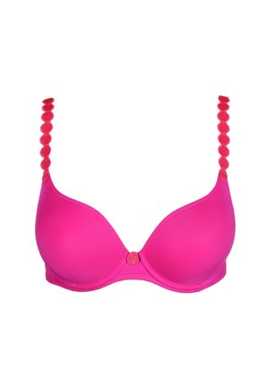 Vrouw poseert in felroze beha met decoratieve bandjes en bijpassende bikini-onderbroek tegen een effen lichte achtergrond.