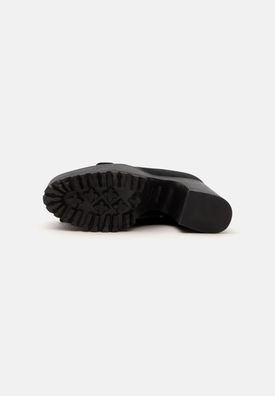 Semelle d'une botte noire avec un talon bloc épais et un motif profond de la bande de roulement, présentée couchée sur le côté sur un fond clair uni.