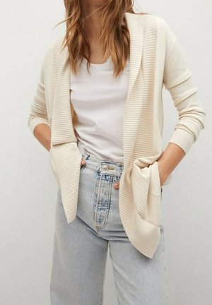 Frau trägt eine cremefarbene, gerippte Strickjacke über einem weißen T-Shirt, das in eine hoch taillierte hellblaue Jeans gesteckt ist, Hände in den Taschen der Strickjacke.