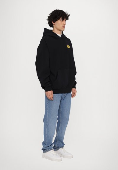 Haut à capuche en coton noir avec une poche avant, logo brodé en jaune, associé à un jean en denim bleu clair et des baskets blanches.