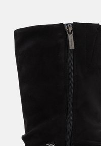 Botín de ante negro hasta el tobillo con cremallera lateral, textura suave y borde superior estructurado. Presenta costuras a lo largo de la línea de la cremallera.