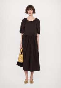 Tumši brūna puff-sleeve krekls ar priekšējiem pogām, apvienots ar plūstošu tumši brūnu svārku. Modelis tur austu dzeltenu somu; uz viņas kājām ir zelta sandalītes.