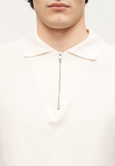 Κοντινό πλάνο ενός ατόμου που φοράει υφασμένο λευκό πόλο με μεταλλικό φερμουάρ στον γιακά, εμφανή χείλη αλλά τα μάτια εκτός κάδρου.