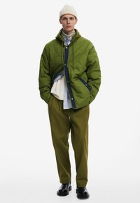 Quilted green padded jacket with navy accents, featuring a hood and snap closure, layered over a white and blue striped shirt and green trousers.