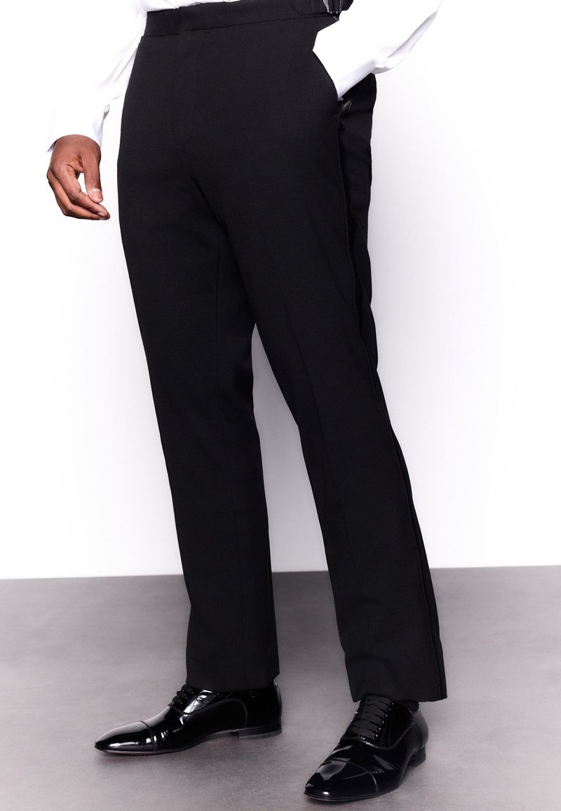 Homme portant un pantalon noir à bande latérale, une chemise blanche et des chaussures de ville noires brillantes, debout sur un sol gris.