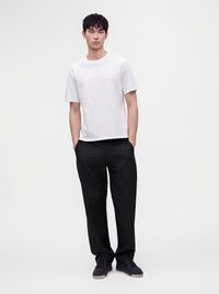 Jeune homme debout, les mains dans les poches, portant un t-shirt blanc à manches courtes, un pantalon noir et des baskets noires sur un fond uni.