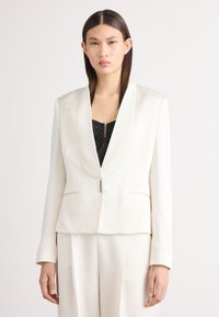 Blazer branco com gola de xale, apresentando um fecho de botão único e dois bolsos frontais, usado sobre uma blusa preta. Textura de tecido suave.