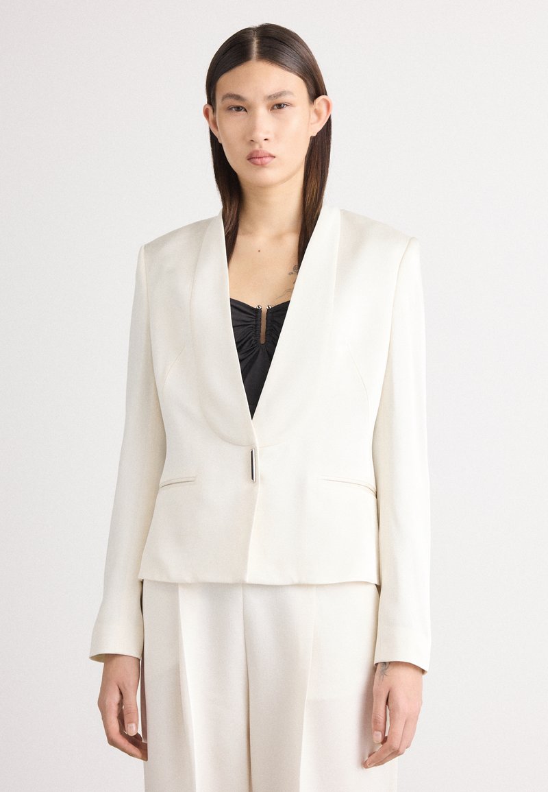 Blazer blanc avec un col châle, comportant une fermeture à un bouton et deux poches avant, porté sur un top noir. Texture de tissu lisse.