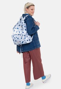 Zaino a pois azzurro chiaro con texture lucida, spallacci regolabili e tasca esterna, abbinato a una giacca di jeans e pantaloni a gamba larga.