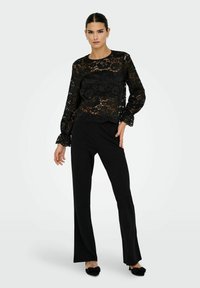 Una donna indossa un top lungo con maniche in pizzo floreale nero, pantaloni larghi neri e tacchi neri con dettagli a fiocco su uno sfondo bianco.