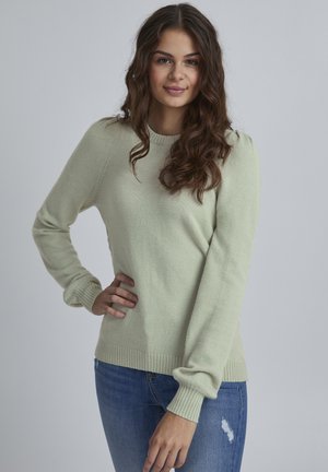 Hellgrüner Pullover aus weichem Stoff, mit Rundhalsausschnitt, langen Ärmeln und gerippten Bündchen und Saum. Getragen mit Jeans aus Denim.