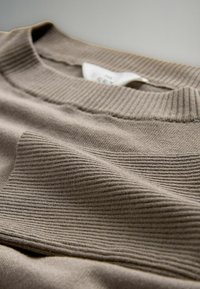 Beige gebreide trui met een geribde kraag en een textuurpatroon; de stof lijkt zacht en heeft subtiele variaties in dikte.