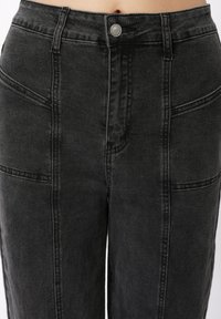 Κοντινή λήψη μαύρου τζιν με ψηλή μέση, κούμπωμα με κουμπιά και εμφανή κάθετα και διαγώνια ραφή στο μπροστινό μέρος.