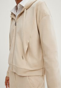Beige zip-up hoodie gemaakt van zacht materiaal, met een capuchon met trekkoord, zijzakken en geribde manchetten. Eenvoudig, minimalistisch ontwerp.