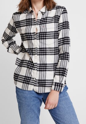 Kvinde iført sort og hvid ternet skjorte med knapper og frontlomme, kombineret med blå jeans, mod en ensfarvet baggrund.