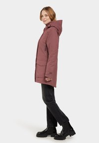 Buitenjack in mauve kleur, met een capuchon, twee zijzakken met rits en een lange pasvorm. Gekozen in combinatie met zwarte jeans en stevige zwarte boots.