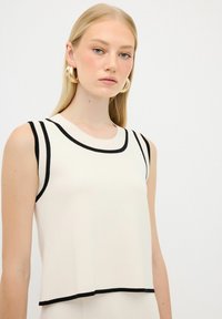 Top senza maniche di colore avorio con accenti in nero a coste lungo il collo e le aperture delle maniche; presenta una vestibilità comoda e un orlo leggermente svasato.