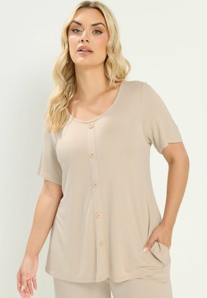 Kvinde med langt blond hår iført en beige kortærmet knap-foran top med en lomme, der står mod en ensfarvet lys baggrund.