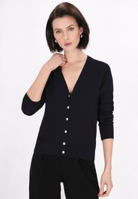 Cardigan blu navy con scollo a V profondo, maniche lunghe e sei bottoni bianchi sul davanti. Realizzato in morbido materiale a maglia texturizzata.