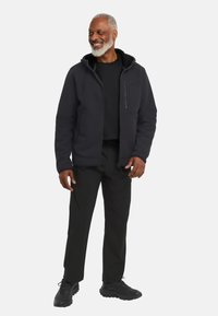 Giacca zip-in grigio scuro con un esterno testurizzato, cappuccio foderato in pile nero e due tasche frontali, abbinata a pantaloni neri e scarpe.