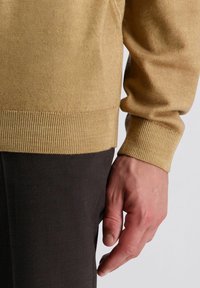 Beige gebreide trui met ribbels bij de mouwen en de zoom, gecombineerd met donkerbruine gestructureerde broek, met een close-up van de hand op de trui.