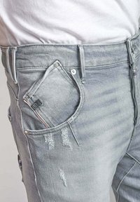 Jasnoszare dżinsy z przetarciami, unikalnym kątem kieszeni i metalowymi akcentami. Widoczna wyblakła struktura.