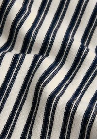 Tissu blanc avec des rayures verticales bleu marine texturées et uniformément espacées, présenté en plis doux créant des ombres délicates.