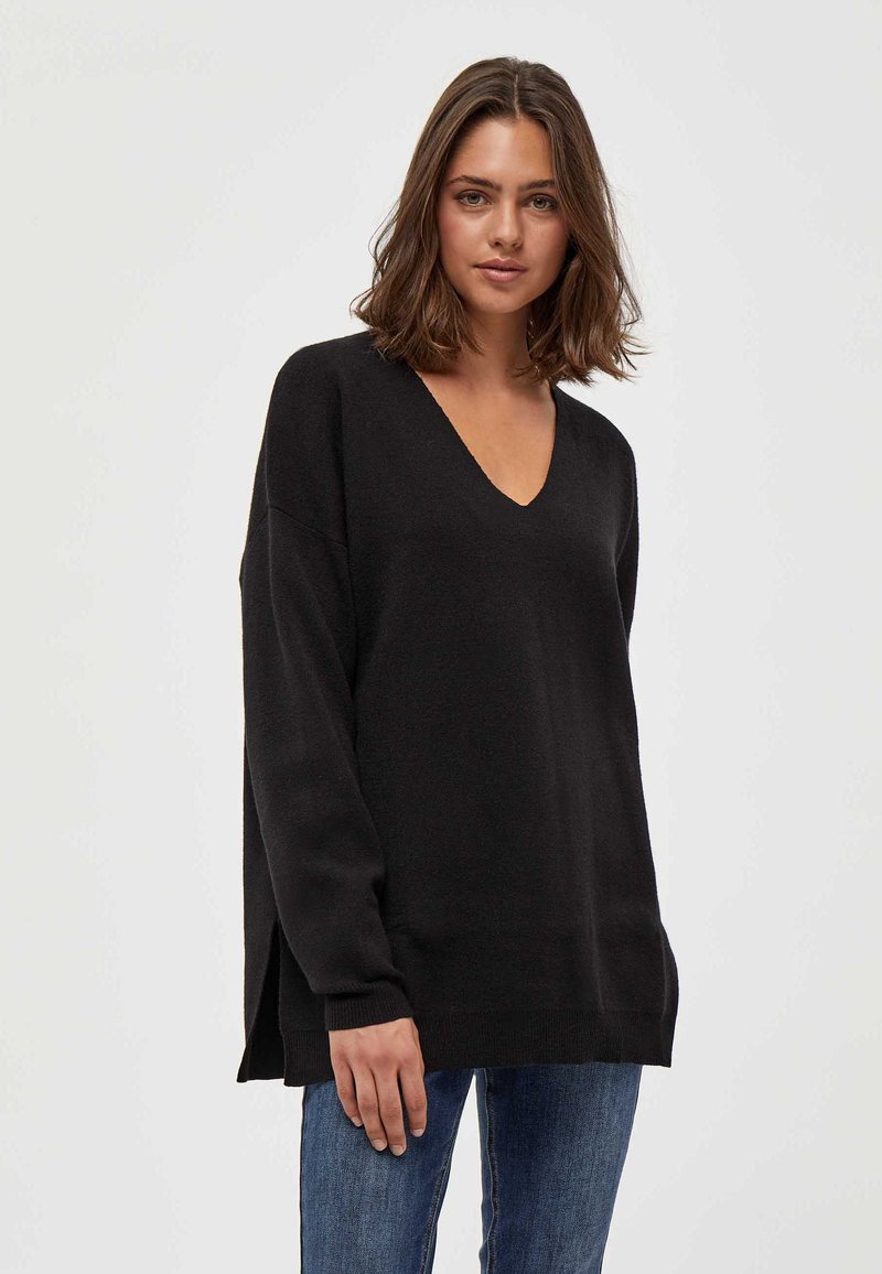 Črn pulover z v-neck iz mehke pletene tkanine, dolgimi rokavi, stranskimi razporki in sproščenim krojem. Ujemajo se z modrimi kavbojkami.