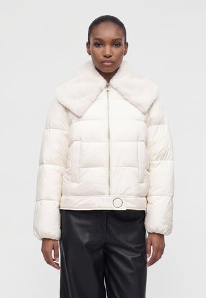 Femme portant une doudoune blanche avec un large col en fourrure et un pantalon noir, debout devant un fond clair uni.