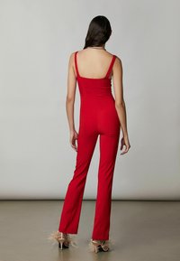Tuta rossa con spalline sottili, corpetto aderente, gambe svasate, texture liscia e design elegante. Le calzature presentano tacchi trasparenti con dettagli in piuma.