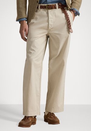 Beige bomuldsbukser med afslappet pasform, udstyret med et flettet brunt læderbælte og brune casual sko. Simpelt design med minimal detaljering.