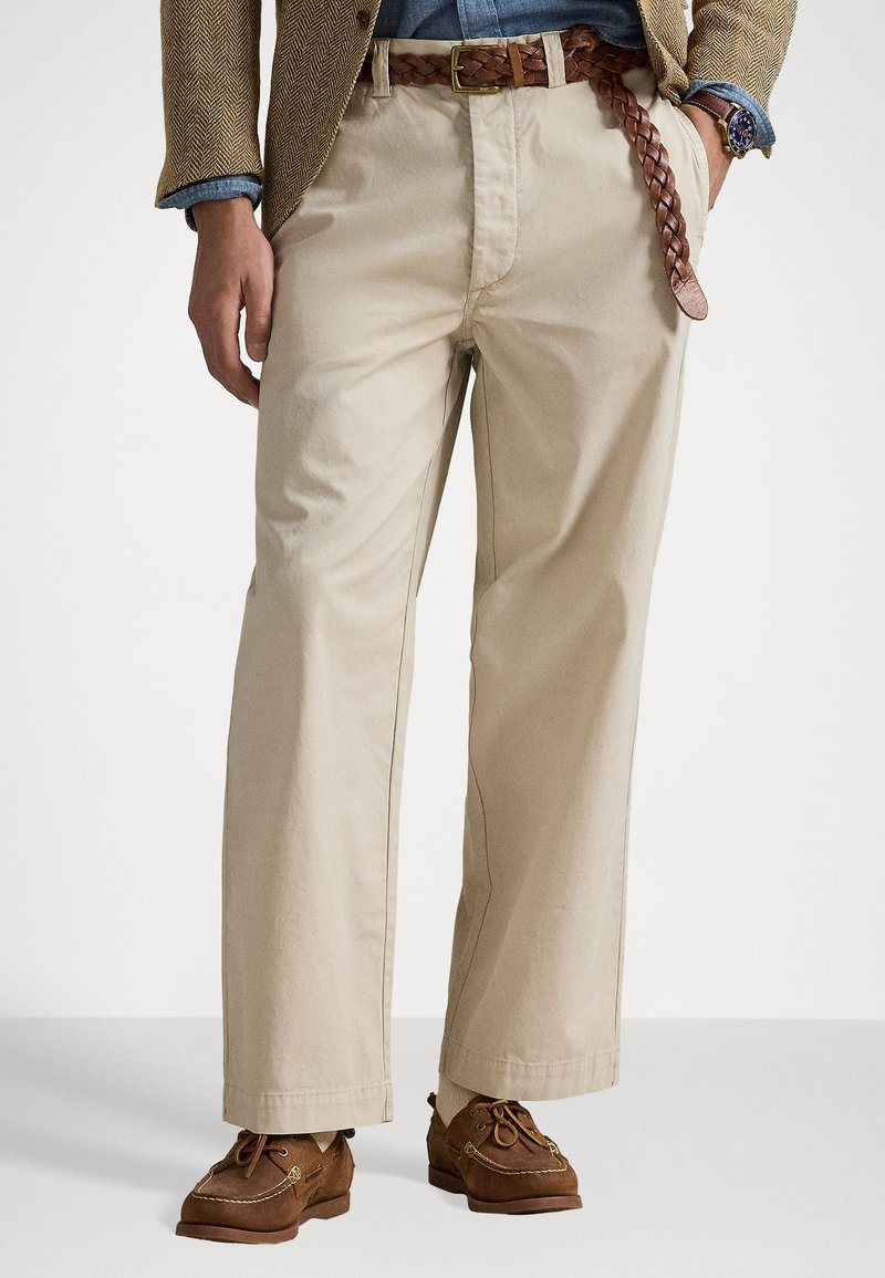 Pantalon en coton beige à coupe décontractée, avec une ceinture en cuir tressé marron et des chaussures décontractées marron. Design simple avec des détails minimaux.