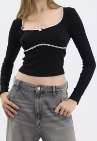 Schwarzes geripptes Langarmshirt mit rechteckigem Ausschnitt und weißer Spitzenborte, kombiniert mit hochgeschnittenen, hellgrauen Jeans.