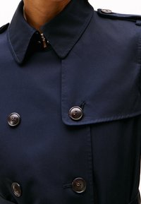 Námořnický trenčkot s dvouřadým zapínáním, velkým límcem a dvěma hnědými knoflíky. Má boční kapsu a hladkou texturu.