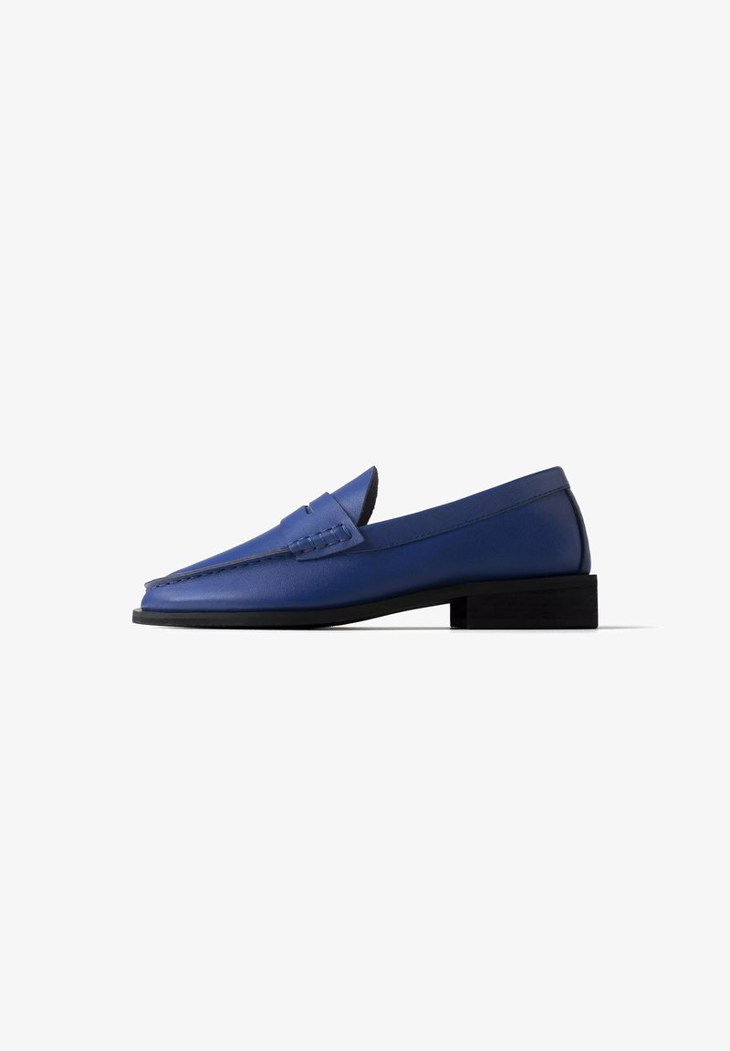 Mocassin bleu en cuir avec petit talon noir et détails cousus sur le dessus, présenté de profil sur fond blanc.