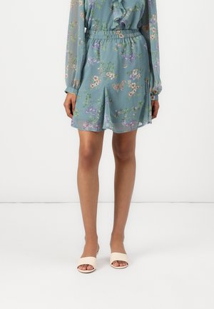 Shorts floraux en tissu vert clair avec une taille élastique. Texture légère avec une coupe décontractée et des fentes latérales. Associés à des claquettes de couleur claire.