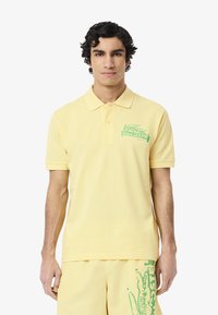 Polo shirt giallo chiaro in cotone, con logo verde Lacoste sul lato sinistro del petto. Maniche corte e design del colletto classico.