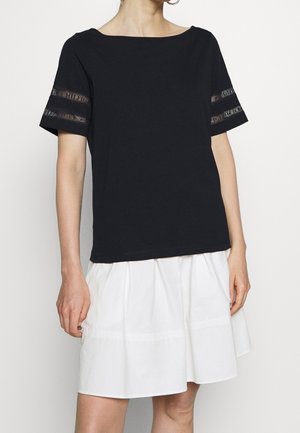 Nainen, jolla on musta, lyhythihainen paita, jossa on läpikuultavat pitsiraidat hihansuissa, ja valkoinen kerroksellinen hame yksiväristä taustaa vasten.