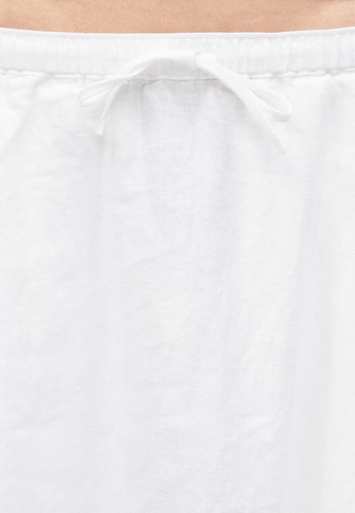 Λευκή υφασμάτινη ζώνη μέσης με δεμένο κορδόνι στη μέση ενός ατόμου, που δείχνει ανοιχτόχρωμο δέρμα πάνω από το ένδυμα.