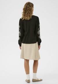 Femme aux cheveux bouclés vue de dos portant un haut à manches longues noir avec de la broderie blanche aux manches et une jupe beige jusqu'aux genoux.