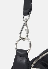 Gros plan d'un mousqueton argenté reliant une sangle en cuir noire à un anneau métallique sur un sac noir à fermeture éclair.