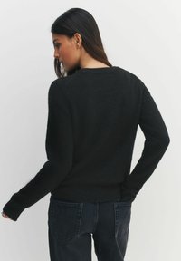 Kobieta z długimi ciemnymi włosami, ubrana w czarny, puszysty sweter i ciemne dżinsy, odwrócona plecami, z jedną ręką w tylnej kieszeni.