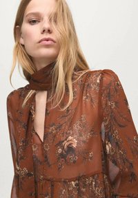 Brązowa bluzka z długim rękawem w kwiatowy wzór, wykonana z przezroczystej tkaniny. Posiada kołnierzyk zapinany na guziki, wycięcie na przodzie i bufiaste ramiona z kwiatowymi wzorami w stonowanych odcieniach.