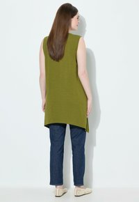Tunic senza maniche verde oliva con orlo curvo, abbinata a jeans blu scuro e scarpe beige senza lacci. Tessuto liscio, design casual.