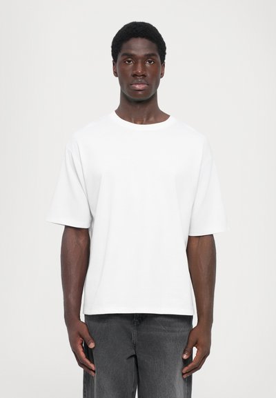 Weißes Baumwoll-T-Shirt mit lockerem Schnitt, rundem Halsausschnitt und kurzen Ärmeln, kombiniert mit dunkelgrauen Jeans. Glatte Textur, minimalistisches Design.